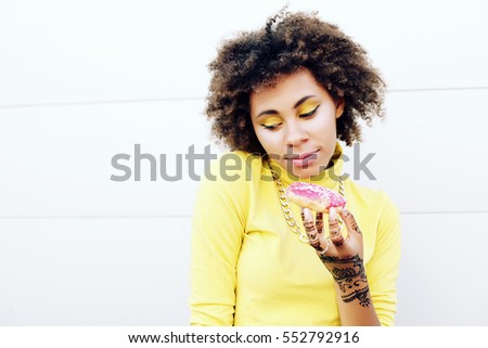Beauty portrait of young african american girl with afro hairstyle. Portrait of hungry woman eating donut on white background. Curly hair, bright art make up. Photo toned style Instagram filters