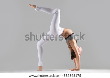 Young attractive woman practicing yoga, standing in Bridge exercise, One legged Wheel pose, working out wearing sportswear, bra, indoor full length, isolated against grey studio background