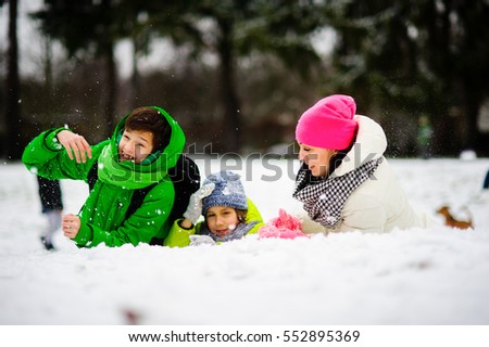 Charming young woman and two of her sons cheerfully spend time in the winter park. They are dressed in bright warm ski suits. All look out because of a big snowdrift.