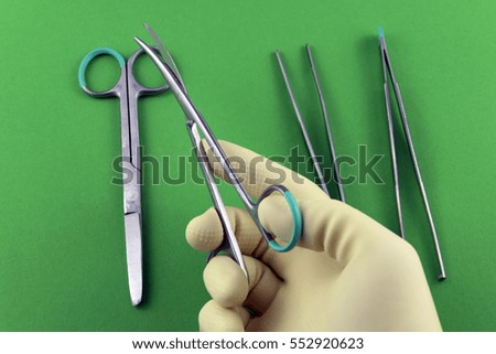 Surgeon hand with sterile glove holding a scissor in front of other surgical instruments aligned as a small surgical set-up and lying on green (pantone) background.