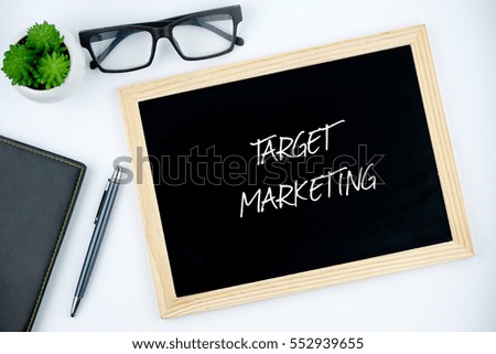 Top view of diary, pen, eye glasses, plant and black chalkboard written with TARGET MARKETING on white background.