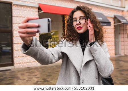 young beautiful woman taking seflie picture using smartphone, autumn street city style, warm coat, glasses, happy, smiling, holding phone in hand, curly hair