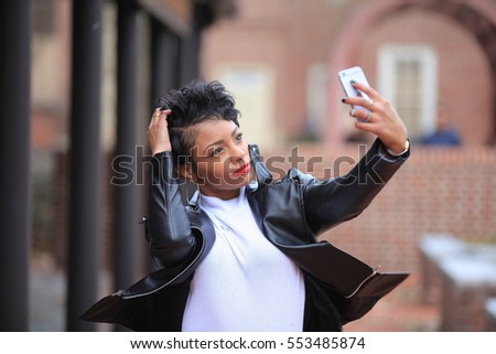 brunette African American woman taking the selfie and smiling with hands near her ears while standing in autumn or spring forest outdoors wearing the black jersey close portrait with a beautiful boke