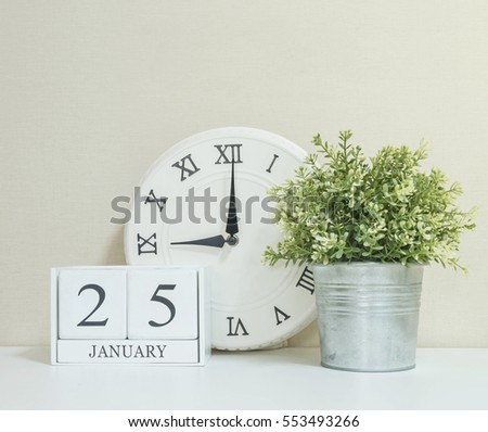 White wooden calendar with black 25 january word with clock and plant on white wood desk and cream wallpaper textured background , selective focus at the calendar