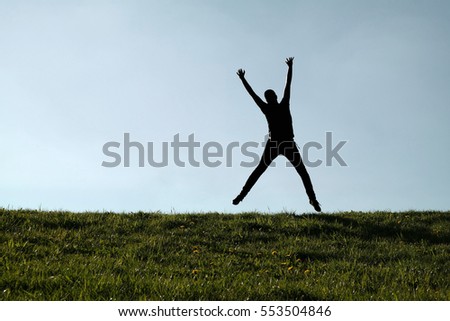 silhouette of single male person jumping high with joyfully raised arms on green grass and blue sky