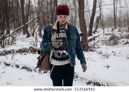 stylish hipster traveler with backpack holding smartphone and browsing in winter snowy forest. wanderlust and adventure concept with space for text. atmospheric moment