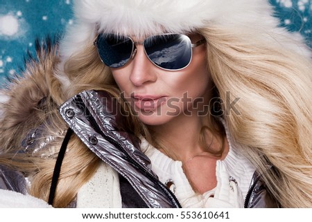 Beautiful stunning woman with long blond hair and perfect face dressed in winter clothing, silver warm jacket and fur cap and silver sunglasses on winter background