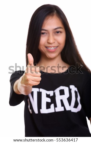 Studio shot of young happy Asian teenage nerd girl smiling and giving thumb up with focus on thumb isolated against white background
