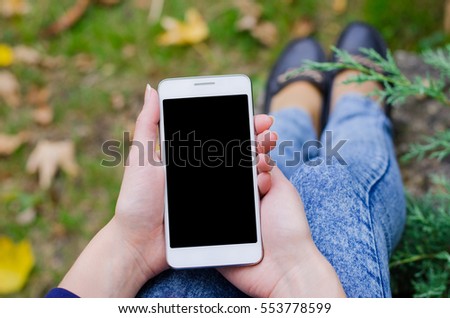 Close up white mobile phone in hand a young hipster business woman in denim blue jeans pants on the background of green natural grass and autumn leaves 