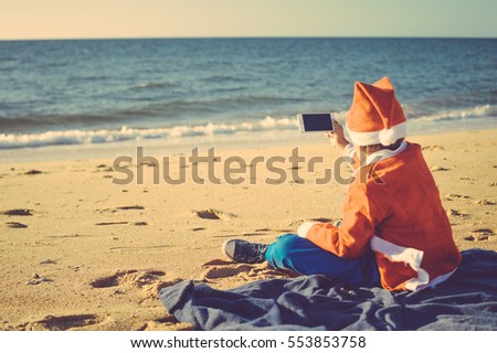 Child boy Santa Claus using mobile phone on ocean beach outdoors background, back view photo. Presents and gifts festive time new year holiday season