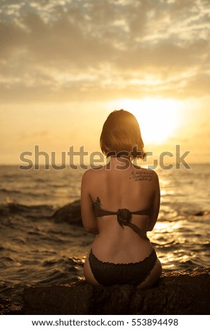 Young girl in bikini sitting on a rock on a seaside. Rising sun on the sky with beautiful woman on first plan.