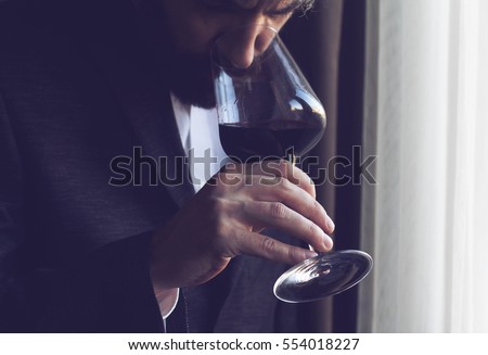 horizontal close up of a Caucasian man with beard, black suit and white shirt tasting a glass of red wine Royalty-Free Stock Photo #554018227