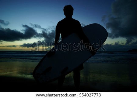 Silhouette of young male surfer standing at the beach and holding surfboard at sunset against blue sky with clouds background.