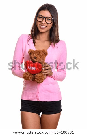 Young happy Brazilian nerd woman smiling while holding teddy bear with heart and love sign isolated against white background