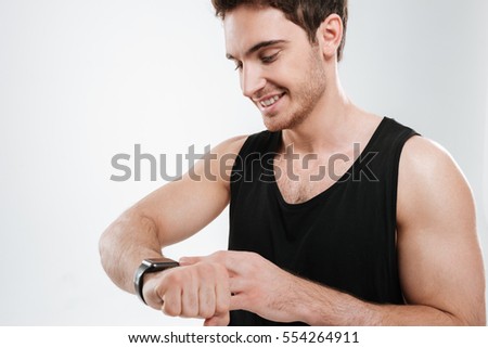 Picture of cheerful young man dressed in black t-shirt standing over white background looking at watch.