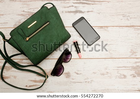 Green ladies handbag, sunglasses, phone and lipstick on wooden background. Fashionable concept, top view Royalty-Free Stock Photo #554272270