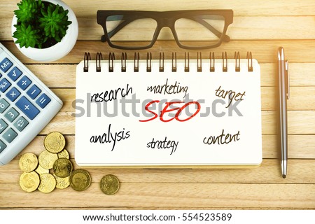 Top view of open notebook written with SEARCH ENGINE OPTIMIZATION (SEO) inscription. Coins, calculator, plant, eye glasses, pen and notebook on wooden background. Business Concept.