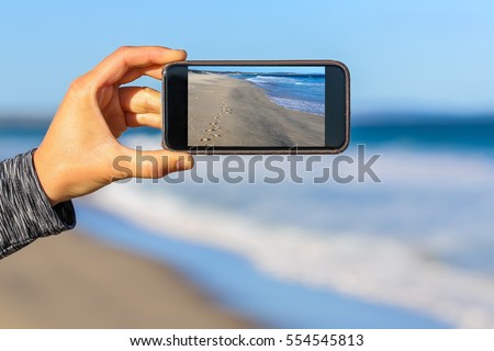 Close up of hand taking a photograph of some footsteps on a beach with on a mobile phone screen