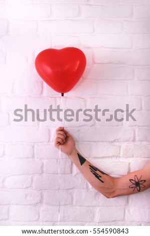 Tattooed hand with red heart balloon on white brick background