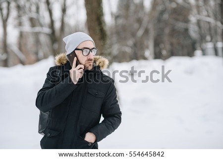 Profile of attractive happy young man with beard, phone standing in winter city. Glasses, jacket, hat, bagpack. Color.