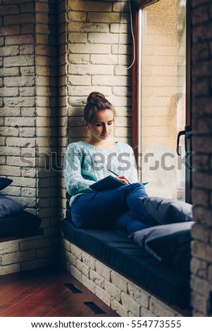 Young beautiful smiling girl in a cyan sweater sitting on the window sill and drawing a picture in sketchbook