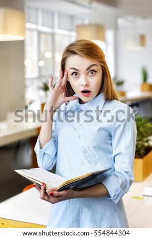 Very surprised pretty office girl with notebook in her hands amazement reacts as she stands in a light office room