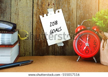 2018 GOALS text written on sticky note. Books, pen, spectacle and red clock on brown desk. Education and business concept.