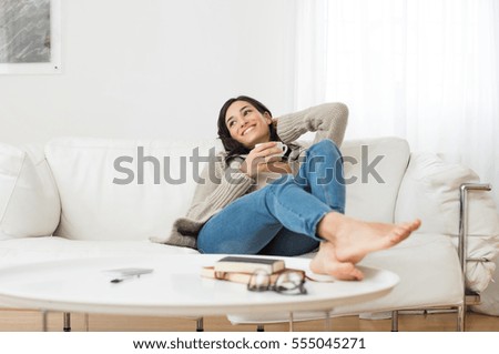 Young smiling woman sitting on sofa and looking up while drinking hot tea. Young brunette woman thinking at home in a leisure time. Happy girl relaxing at home on a bright winter morning. Royalty-Free Stock Photo #555045271