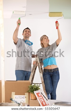 Portrait of smiling couple standing on ladder, painting ceiling of new home with paint roller.
