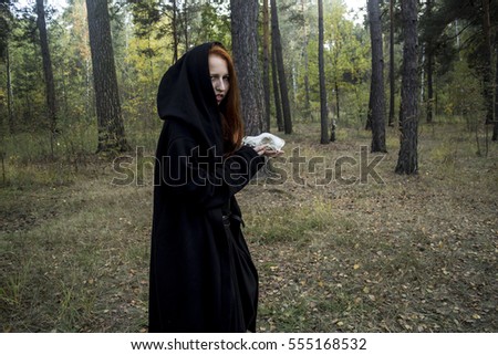 Young beautiful red-haired girl dressed like a witch in a black gown and dark clothes, with long hair, looks like a fox in the autumn forest