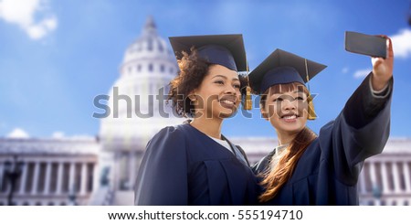 education, graduation, technology and people concept - group of happy international students in mortar boards and bachelor gowns taking selfie by smartphone over american white house background