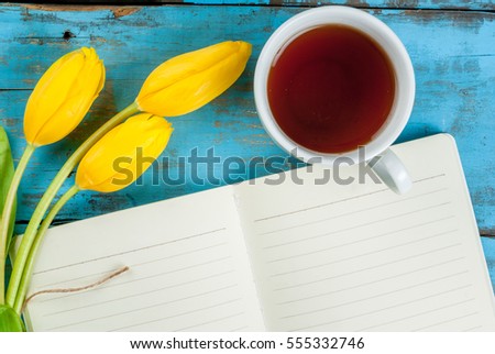 Tea mug with flowers yellow tulips and notebook for notes or wishing on blue rustic table top view, copy space, for Valentine's, Mothers day or Women's day