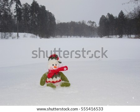 Snowman isolated sits on the snow surface. Snowman has green, red and white clothes. Background consists with snow capped trees. Snow background, winter landscape.