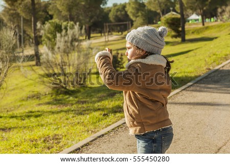 Little girl wearing grey bonnet and jeans taking pictures with a smartphone in the park