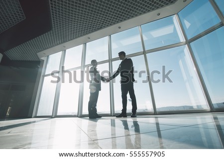 Two young businessmen are shaking hands with each other standing against panoramic windows. Royalty-Free Stock Photo #555557905