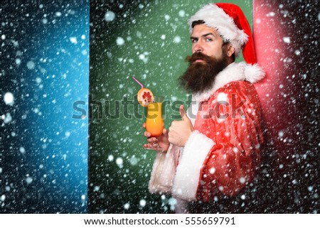 handsome bearded santa claus man with long beard on serious face holding glass of nonalcoholic cocktail in christmas or xmas sweater and new year hat on colorful studio background,copy space