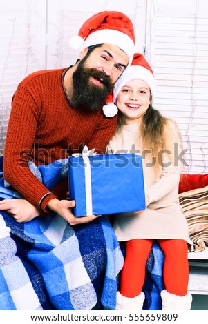 handsome bearded man with long beard in christmas red hat with blue checkered plaid holding big present and cute blonde offended girl in beige dress with crossed hands on white studio background