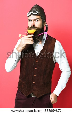 handsome bearded pilot or aviator man with long beard and mustache on serious face holding glass of alcoholic beverage in vintage suede leather waistcoat with hat and glasses on red studio background