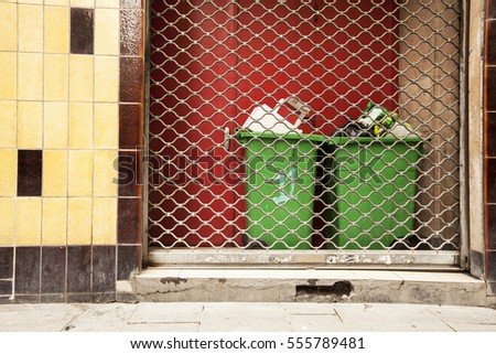 Two green dumpsters behind metal roll bar in front of red door, next to a yellow tile wall, back alley or passage, Paris France