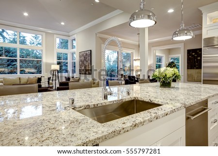 White kitchen design features large bar style kitchen island with granite countertop illuminated by modern pendant lights. Northwest, USA Royalty-Free Stock Photo #555797281