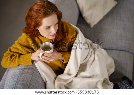 Relaxed young redhead woman enjoying a tea break sitting wrapped in a warm blanket on a comfortable couch staring thoughtfully ahead, high angle view Royalty-Free Stock Photo #555830173