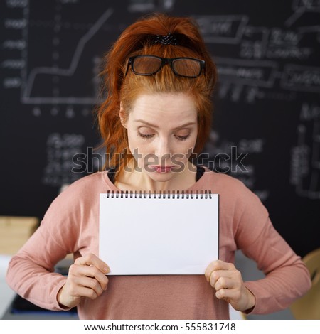Young red haired woman sadly looking down on blank sheet of paper she holding in hands, standing in classroom or office