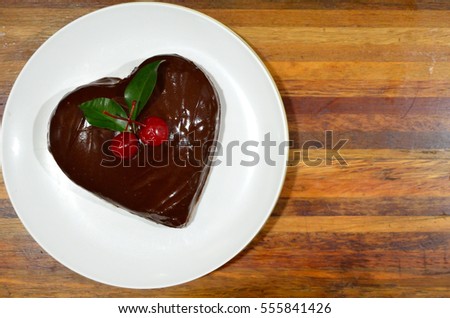 Heart shape homemade chocolate cake is decorated by cherry topping and wooden background.
