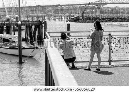 Asian couple romantically placing a love lock onto a gate full of padlocks by the marina with a vintage boat and harbor in the background in street photography style black and white