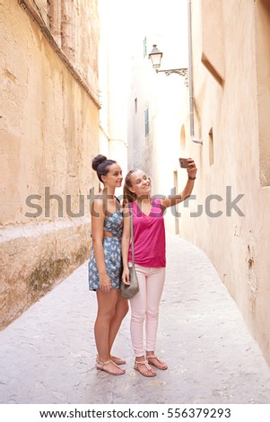 Two smiling teenagers girls friends visiting a destination city street with stone buildings together, taking selfies pictures on student trip holiday, sunny outdoors. Travel technology lifestyle.