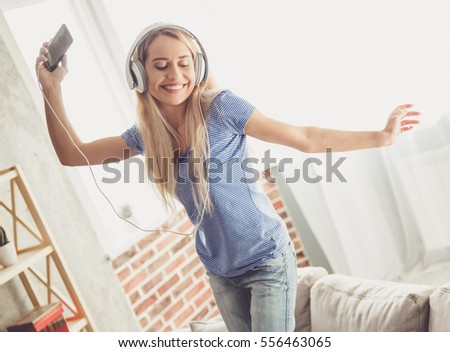 Beautiful girl in headphones is listening to music using a smartphone, smiling and dancing at home