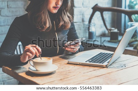 Young woman sitting in coffee shop at wooden table, drinking coffee and using smartphone.On table is laptop. Girl browsing internet, chatting, blogging. Female holding phone and looking on his screen. Royalty-Free Stock Photo #556463383