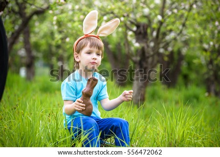 Happy little toddler boy eating chocolate and wearing Easter bunny ears, sitting in blooming garden on warm sunny day. Celebrating Easter traditional holiday.