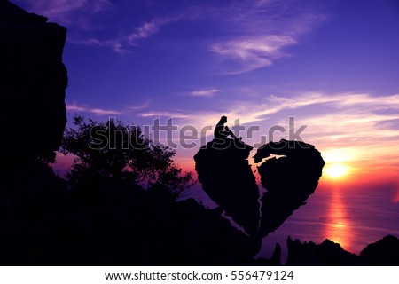 Women sit on broken heart-shaped stone on a mountain with purple sky sunset background.Silhouette Valentine background concept. Royalty-Free Stock Photo #556479124