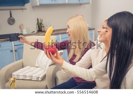 Toned picture of friends girls posing for mobile or smart phone's camera with different fruits while having party at home. Home party concept.